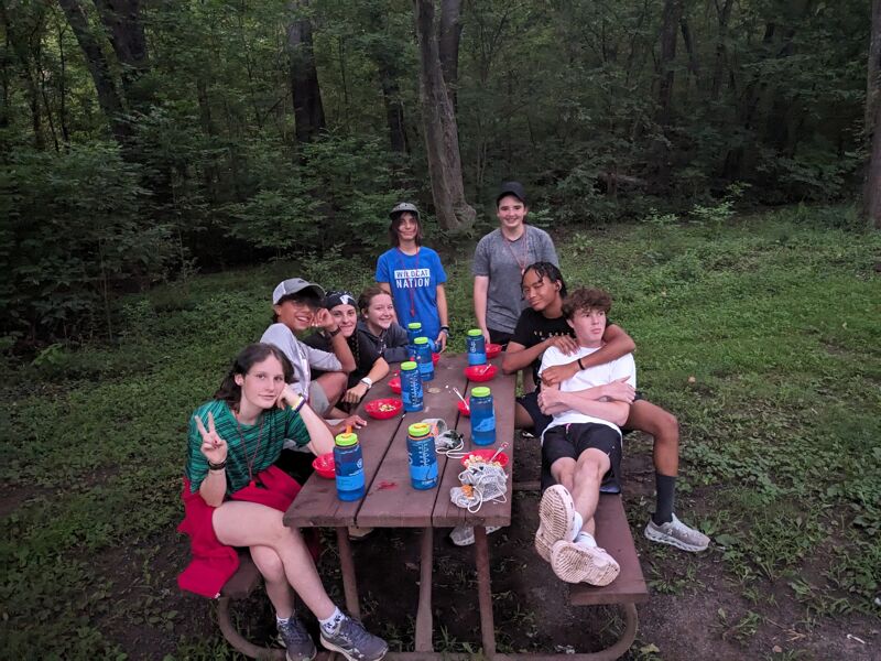 A group of about ten young people are gathered around a wooden picnic table in a wooded area. They appear to be on an outing or camping trip, as suggested by the natural setting. Some of them are sitting on the benches, while others are standing or leaning against the table. There are water bottles on the table. They seem to be enjoying each other's company.